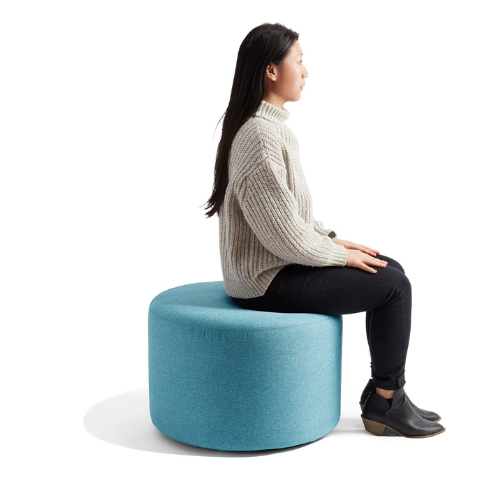 Woman sitting on blue ottoman in white sweater and black pants against white background. (Blue)