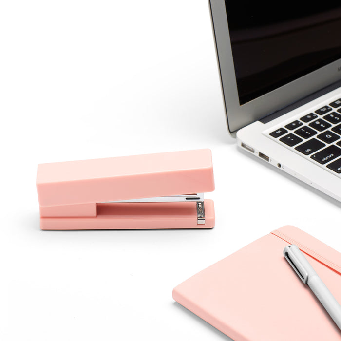 Pink stapler on desk next to open laptop and notebook with pen. (Blush)