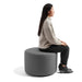 Woman sitting in profile on a round gray ottoman against a white background. (Dark Gray)