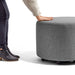 Person leaning on a round gray fabric ottoman with wheels on a white background. (Dark Gray)