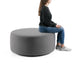 Woman sitting on a large round gray ottoman on a white background. (Gray)
