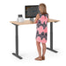 Woman standing at adjustable standing desk working on computer in a home office. (Natural Oak-47&quot;)