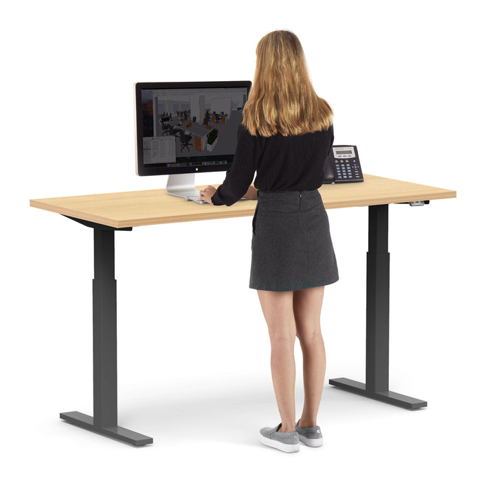 Woman standing at an adjustable height desk working on a desktop computer. (Natural Oak-60&quot;)