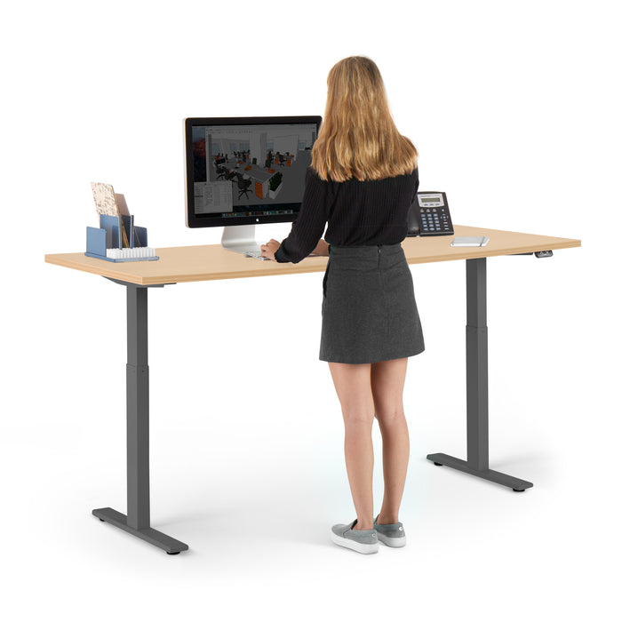 Woman standing at a modern height-adjustable office desk working on a computer. (Natural Oak-72&quot;)