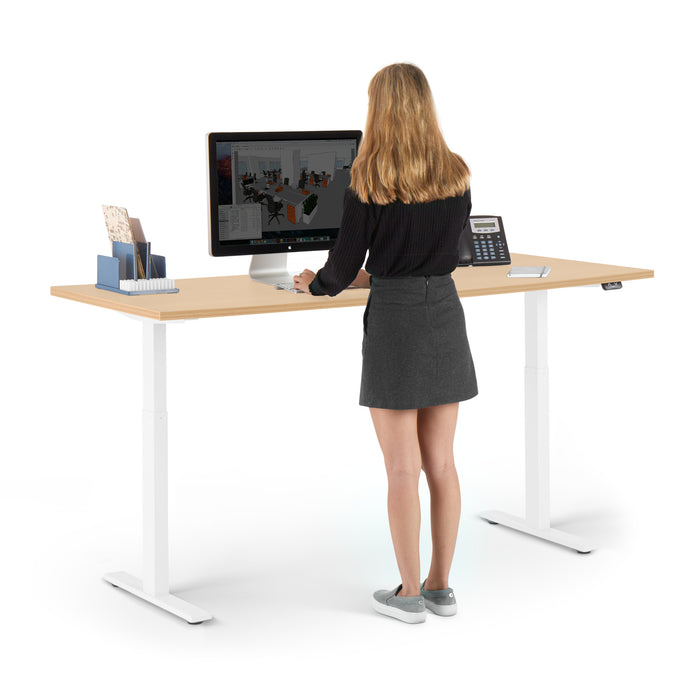 Woman standing at an adjustable desk working on a desktop computer in an office setting. (Natural Oak-72&quot;)