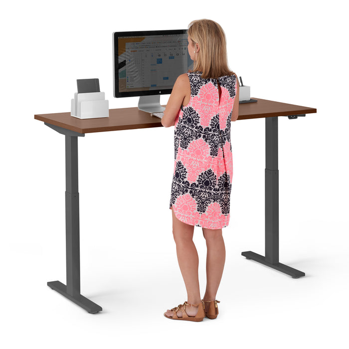 Woman standing at adjustable standing desk working on computer in office setting. (Walnut-57&quot;)