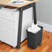Slim trash can with crumpled paper beside white office drawer unit and desk on wooden floor. (Charcoal)