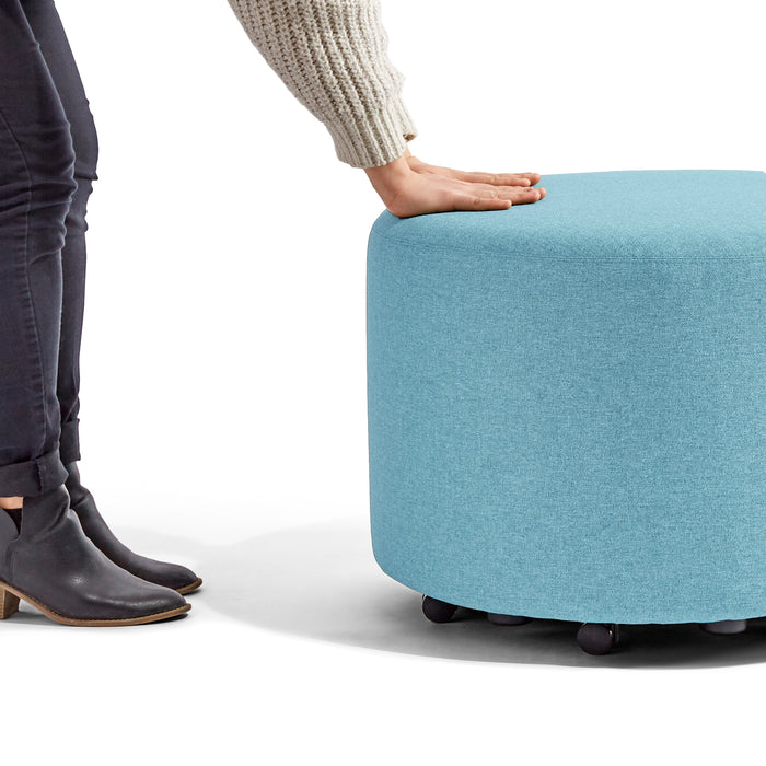 Person reaching out to touch a blue round fabric ottoman with wheels on a white background. (Blue)
