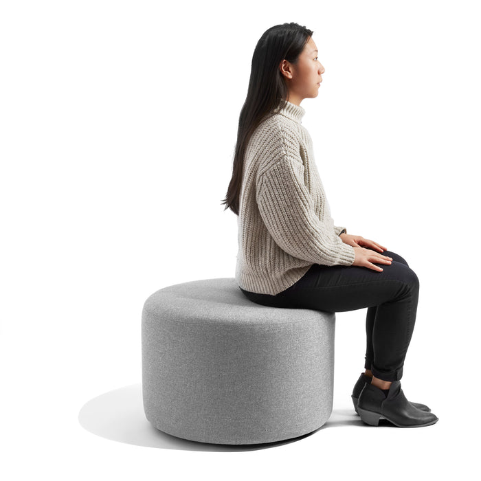 Woman in a beige sweater sitting on gray modern ottoman with white background. (Gray)