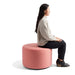 Side profile of a woman sitting thoughtfully on a round pink ottoman against a white background. (Rose)