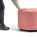 Person reaching out to touch a round pink ottoman on wheels against a white background. (Rose)