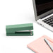 Green stapler next to silver laptop and pink notebook with pen on white background. (Sage)