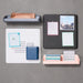 Flat lay of organized desk accessories with notepads, stationery, and a passport on a grey background. 